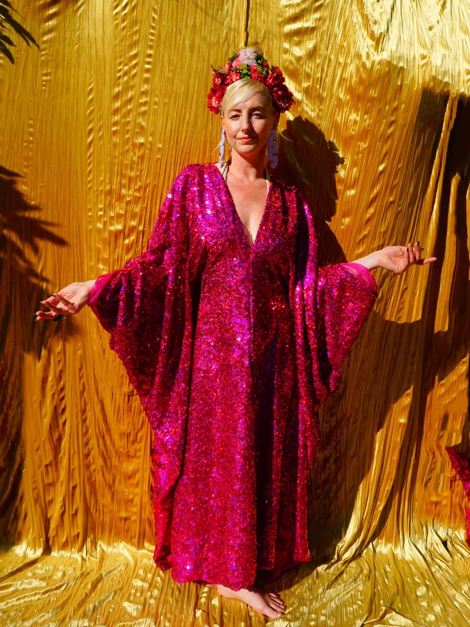 Cerise holographic sequin kaftan seen on a woman standing in front of a gold background with her hands in the hair.
