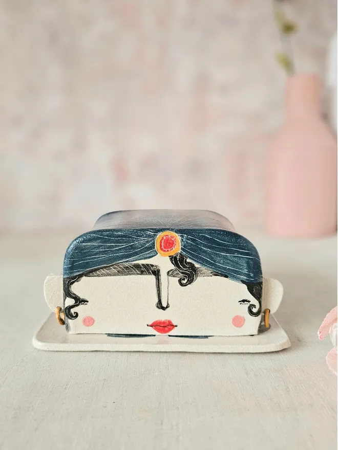 A whimsical ceramic butter dish shaped like the head of a fortune teller, painted in an illustrative style. The fortune teller wears a turban with a few curls escaping, and has a mystical expression. The base of the dish features delicate flowers, an all-seeing eye, and the playful words “I predict you need to buy some butter,” adding a quirky touch of fortune-telling flair to your kitchen.