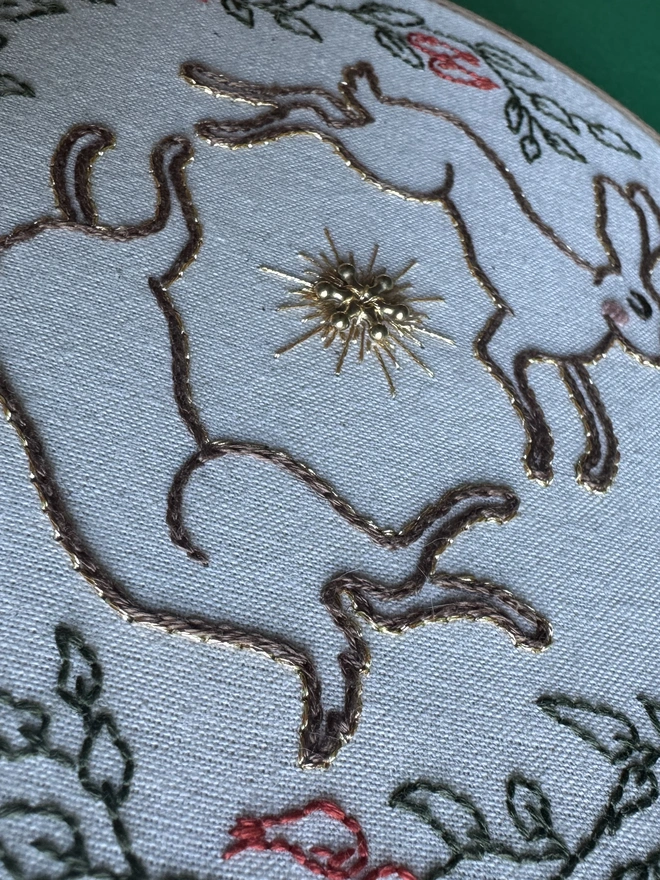 Embroidered hares edged in gold circling a gold bead and gold floss