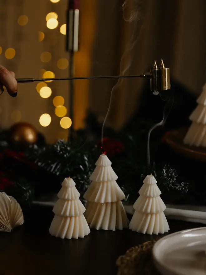 White Christmas Tree Warm Candles (Set Of 2). Three unlit cream tree candles in graduating heights on a dark table, with frosted pine garland, baubles and golden bokeh lights behind. The fluted design gives a soft, Scandi-inspired look to holiday tablescapes.