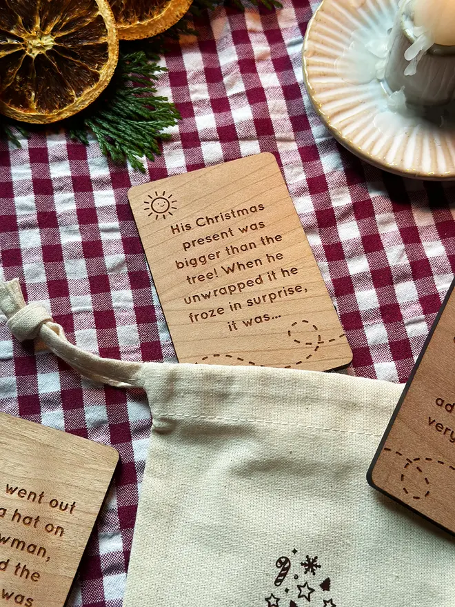 Close-up of engraved wooden Christmas story prompt cards on a red gingham cloth beside a pink book, showing a mix of story beginnings and endings for children’s festive storytelling.