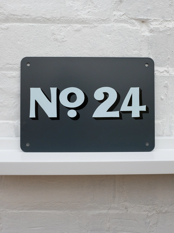 Hand painted house number in flourish style, pale blue and black No. 24 on anthracite grey metal plaque, against a white brick wall.