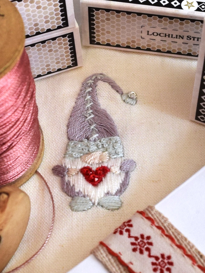 A mini embroidered Christmas Gonk in winter blue and lilac holding a big warm scarlet heart that twinkles with crystals.