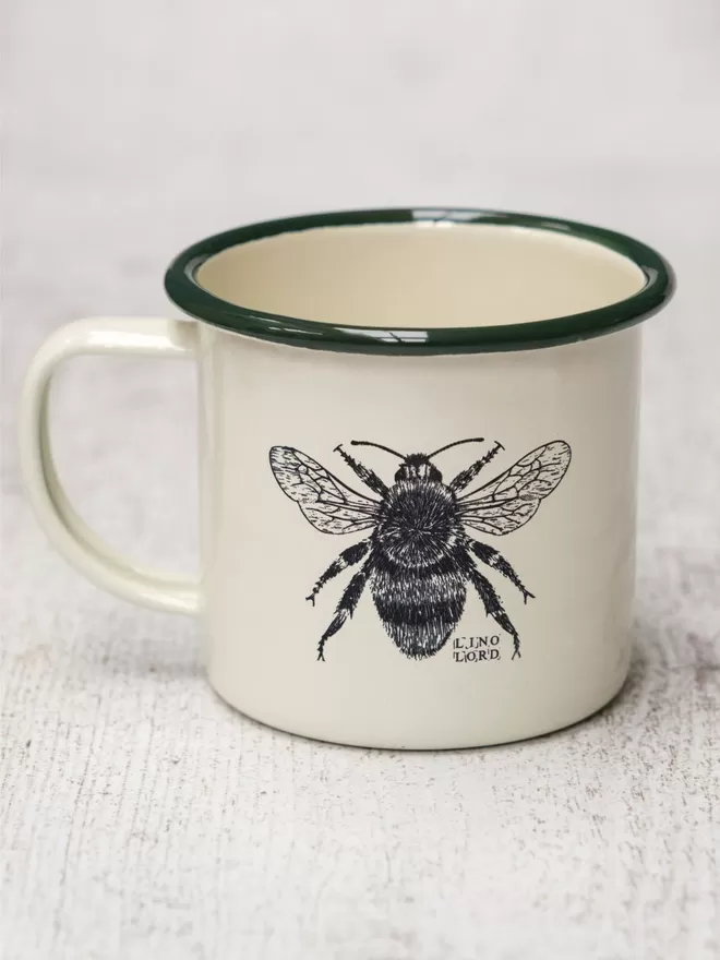 Picture of a Cream Enamel Mug with a Green Rim with a Bee design etched onto it, taken from an original Lino Print