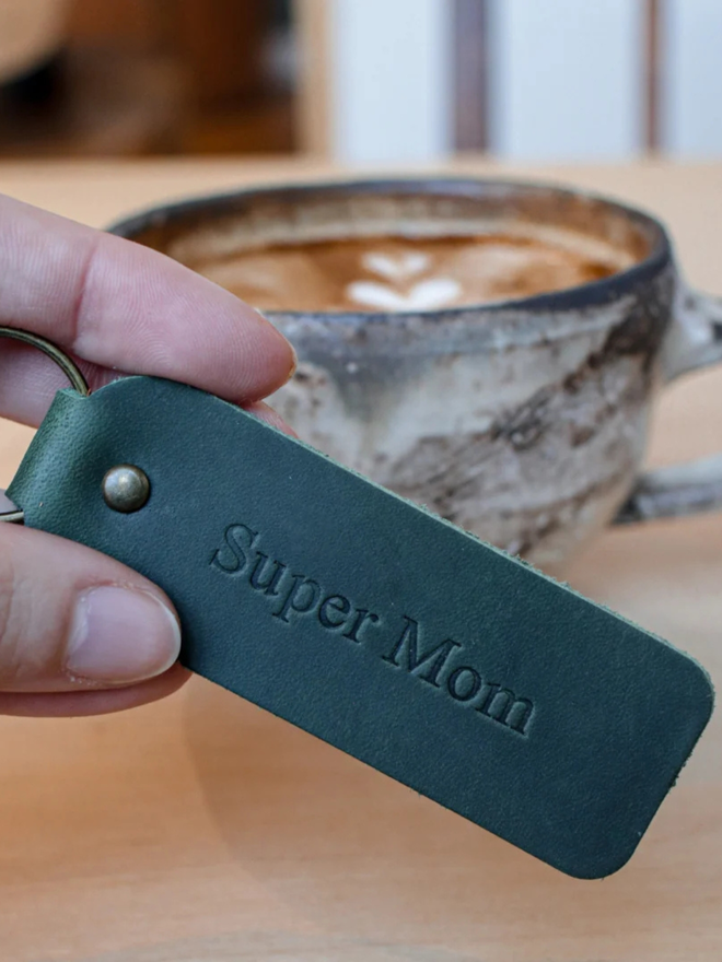mother's day leather keyring. a hand holding a green keyring embossed with 'super mom'.