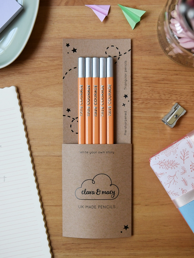 Five orange pencils with the words Have Courage along the side of each one, are tucked into cardboard packaging on a wooden desk.