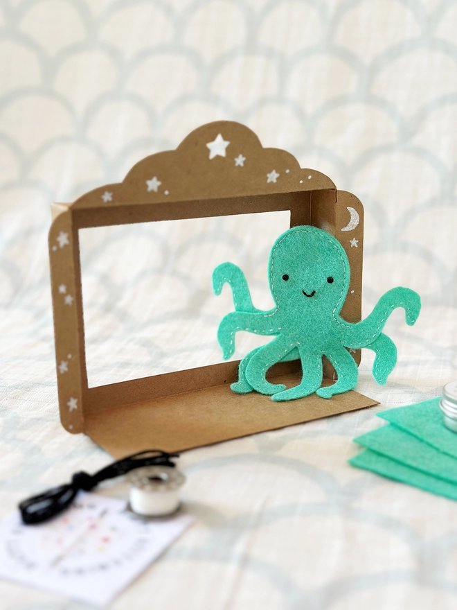 A handmade mini cardboard puppet theatre stands on a pale fabric surface, with a turquoise octopus finger puppet beside it.