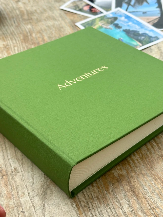 a green linen photo album is on a wooden coffee table, There are some photos in the background of beautiful landscapes.