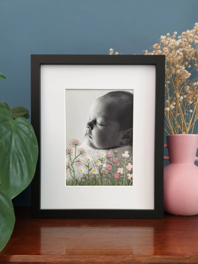 Baby photo with hand embroidered flowers growing from bottom of image, black frame on desk