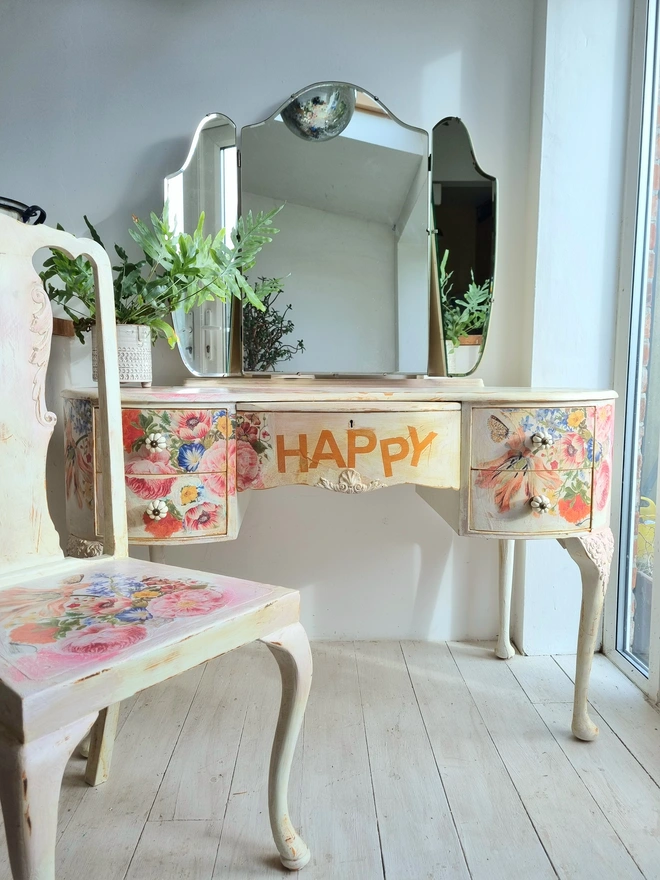 An antique kidney-shaped dressing table with chair and a trio of mirrors. Upcycled in cream and pale pink with decoupage florals in oranges and pinks. Text across the table read ‘flowers make me happy’ in the orange of the undercoat.