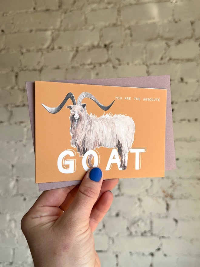 an orangey yellow greetings card featuring an illustration of a bilberry goat with the text “you are the absolute GOAT” with a muted purple envelope