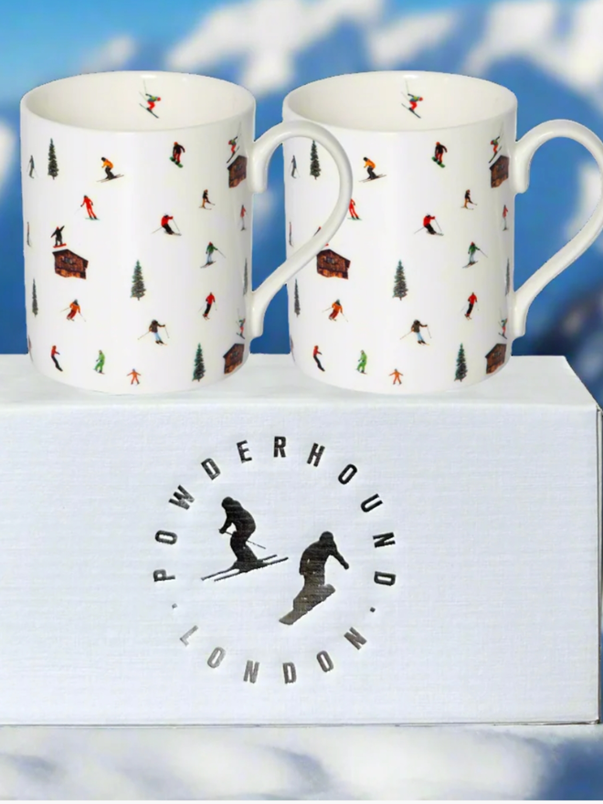 Skit Hut Mug. A pair of white mugs set on top of a gift box with the Powderhound logo. The design is a pattern of multicoloured skiers, ski huts, and trees.