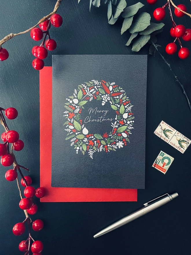 Christmas Card with Pressed Winter Leaf Wreath Design Made from Pressed Winter Leaves, Holly, and Ivy on Red Envelope - Dark Charcoal Desk Setting