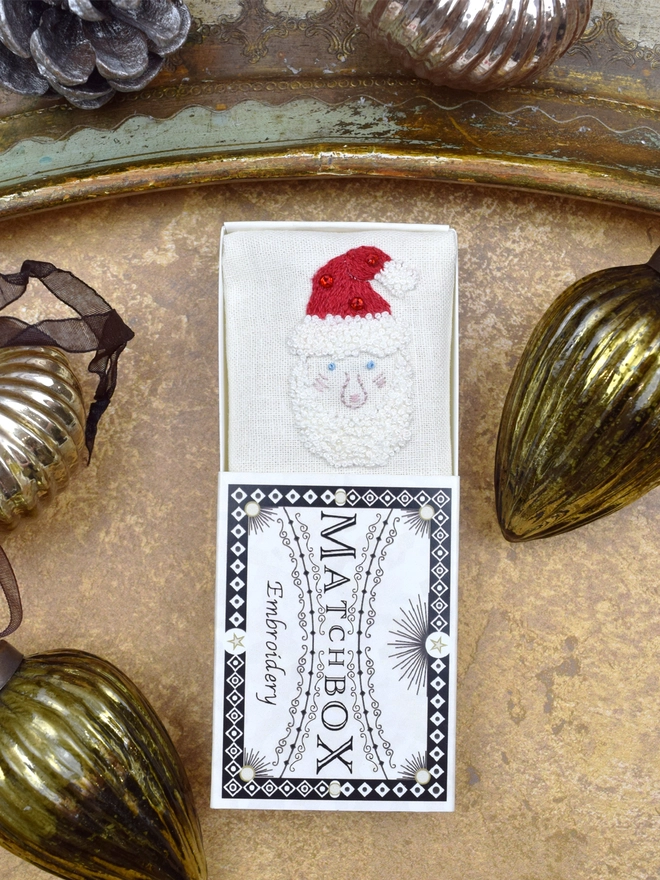 An embroidered Santa, inside a Matchbox displayed on a gold background with glass baubles.