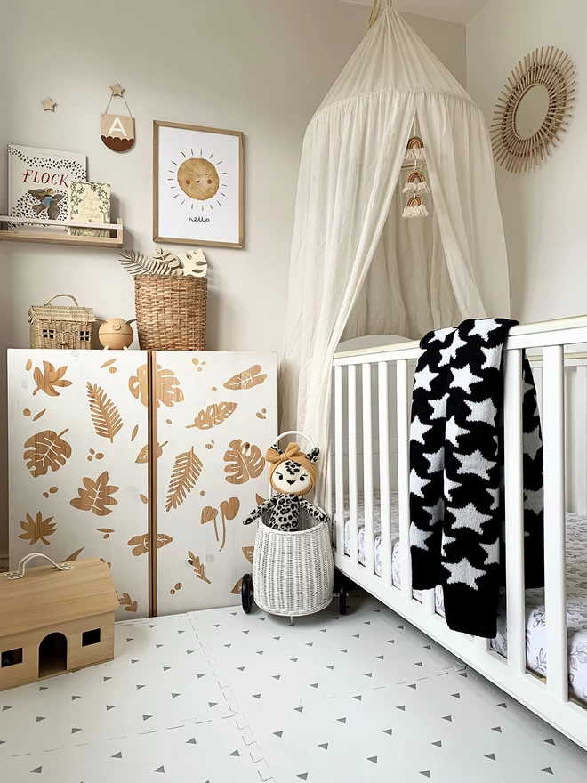 A neutral scandi style nursery with lots of natural wood, wicker accessories and a white cot. Over the cot is draped a star baby blanket with a black trim. showing the reverse colourway, white stars on black.
