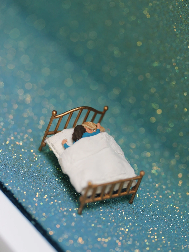 Miniature scene in an artbox showing two men lying in bed against a sparkling backdrop evoking the night sky.