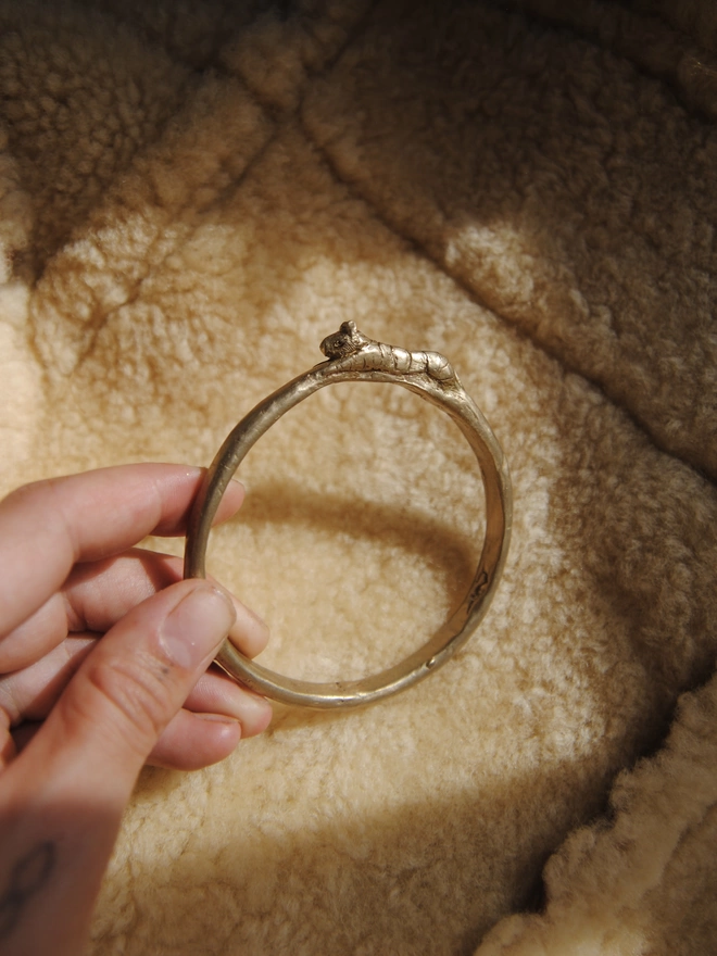 The image of a hand holding a gold toned brass bangle with a hand-carved tiger on it, the background of the image is a beige sheepskin fleece 