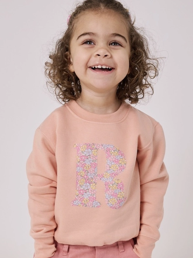 a 2 year old girl wearing a personalised dusty pink sweatshirt with her initial sewn on the front in floral liberty print 