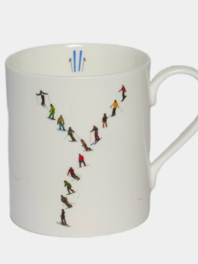 A simple white mug illustrated with a ‘Y’ made of small, multicoloured skiers. The mug is set against a plain, white background.