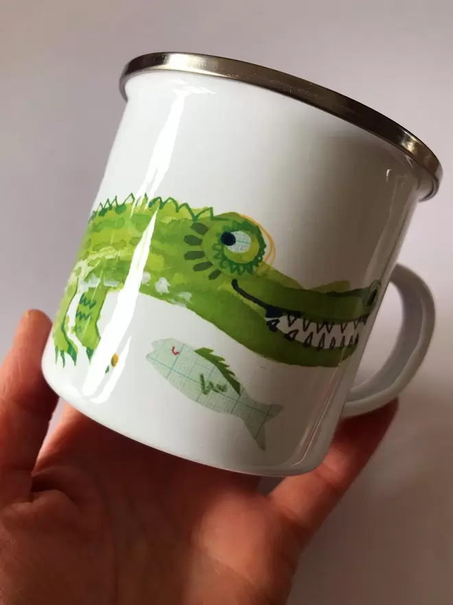 Hand holding a white shiny enamel mug decorated with a bright green crocodile deisgn