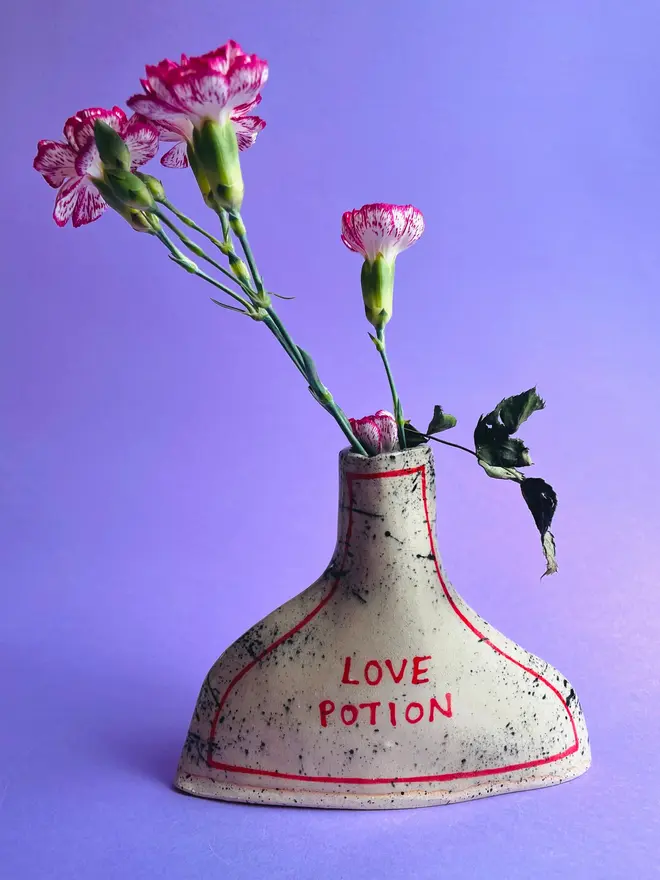 Love Potion/ Poison Bottle Vase, a vase that says 'love potion' in front of a purple backdrop. Each is stamped with a signature red dot on the bottom. 