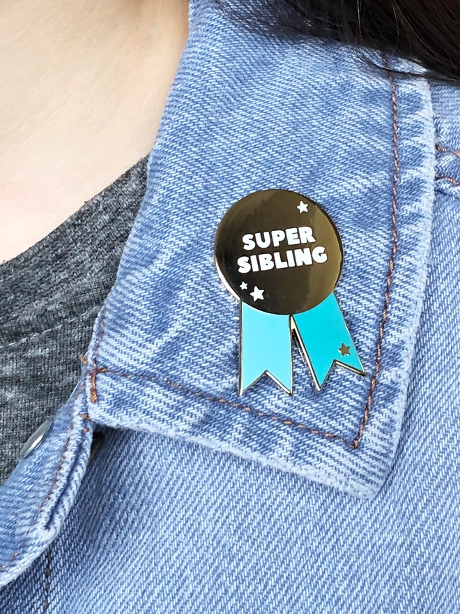 A turquoise and gold pin badge in the shape of a rosette is pinned to the collar of a denim jacket. It has the words "Super sibling" on.
