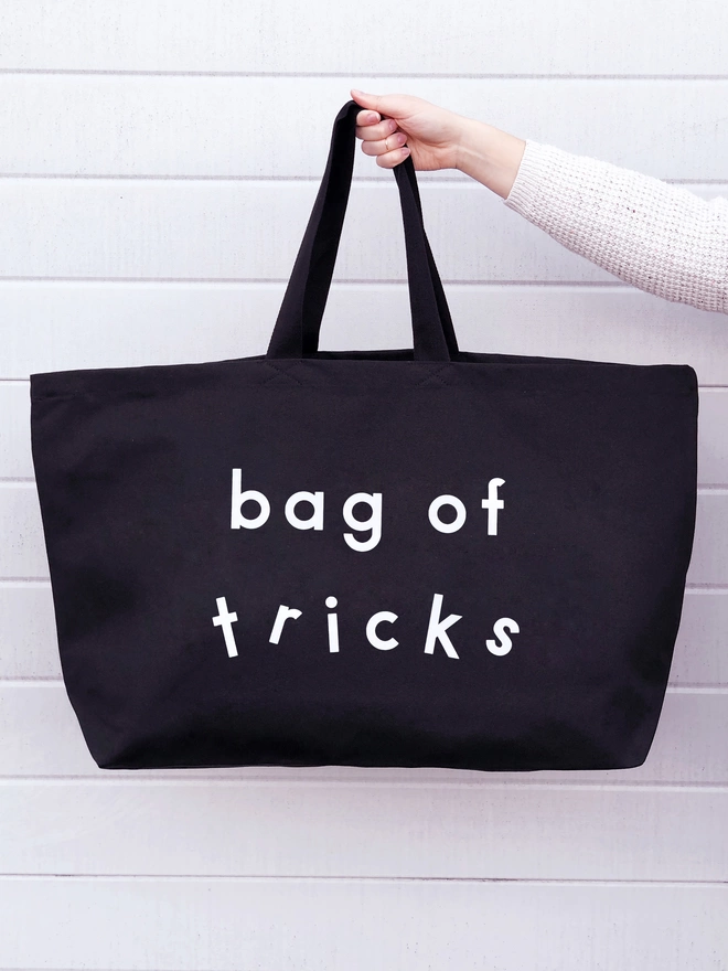 a hand holding out model holding out an oversized black canvas tote bag with the words bag of tricks