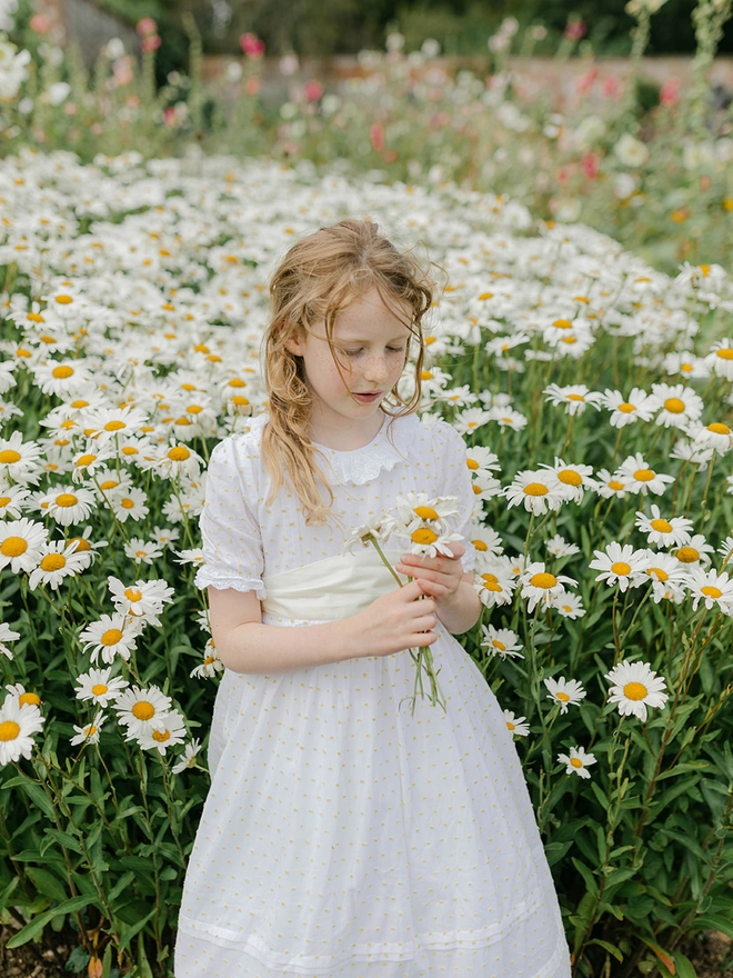 A girl in a while dress with yellow swiss dots and a yellow sash is surrounded by large daisies.