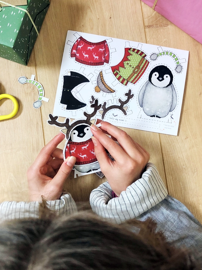 A child is playing with a dress up penguin paper doll that has been cut from a Christmas greetings card.