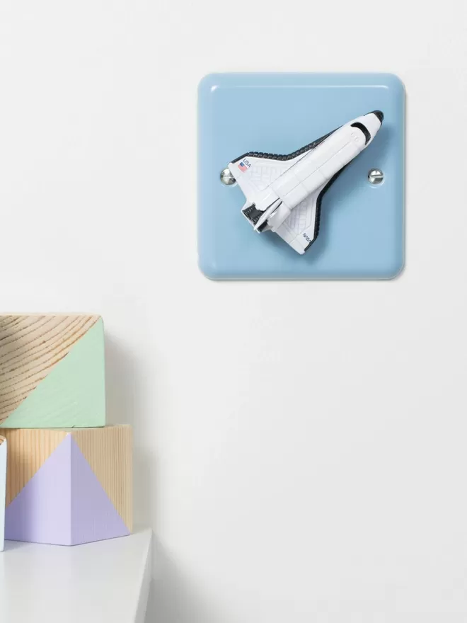 A pastel blue children's space themed dimmer light switch with a white space shuttle as the rotary knob to turn the lights on and off on a white wall next to a white shelf with two wooden nursery building blocks sitting on it. The light switch plate is pale blue and made of metal, epoxy coated steel by Varilight. The space rocket is made of metal. The children's light switch brand is Candy Queen Designs.