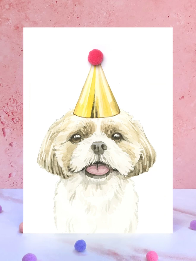 A greeting card featuring a hand painted design of a shih-tzu, stood upright on a marble surface surrounded by pompoms. 