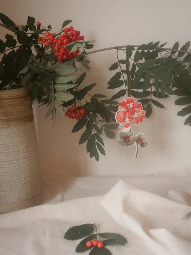 Handmade embroidered geranium Christmas decoration styled with green seasonal foliage and red berries.