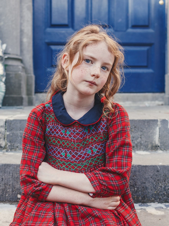 A girl sits on stone steps wearing a red tartan dress with smocking and a navy collar