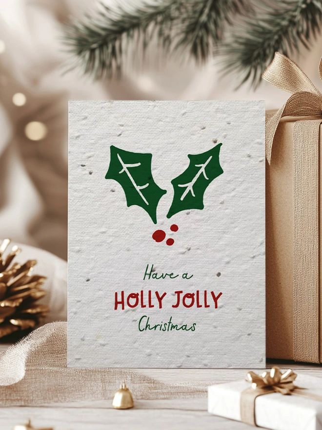 Christmas card with a holly leaf on with 'Have a Holly Jolly Christmas'. Behind the card is some presents, a pine cone and pine leaves.