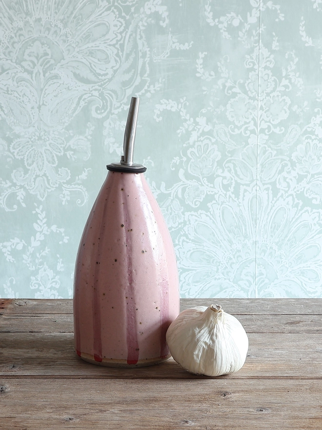 Handmade Ceramic Stoneware Oil Pourer in Pink Stripe sat on a wooden surface next to a bulb of garlic. 