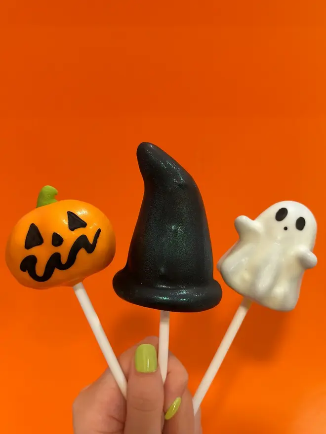 Set of 3 cake pops - one pumpkin, one ghost and one black witches hat, held by a human hand with bright green nails, against a bright orange background