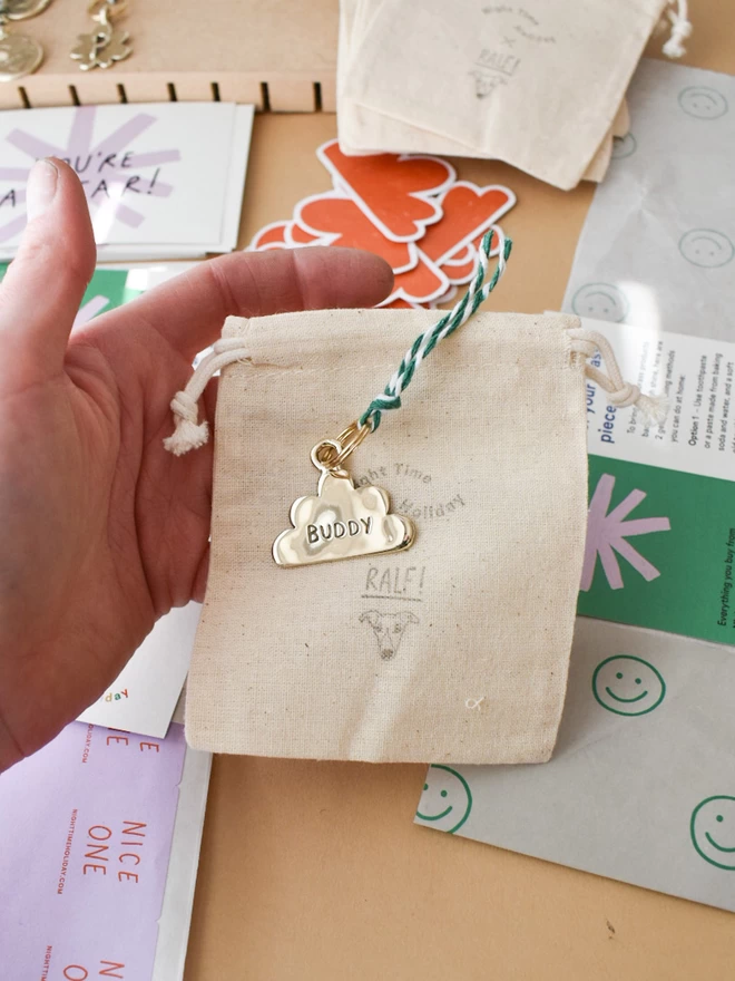 a finished pet tag waiting to be packed, in the shape of a cloud on a cotton drawstring bag. in the background is colourful branded packaging ready to be put into each parcel.