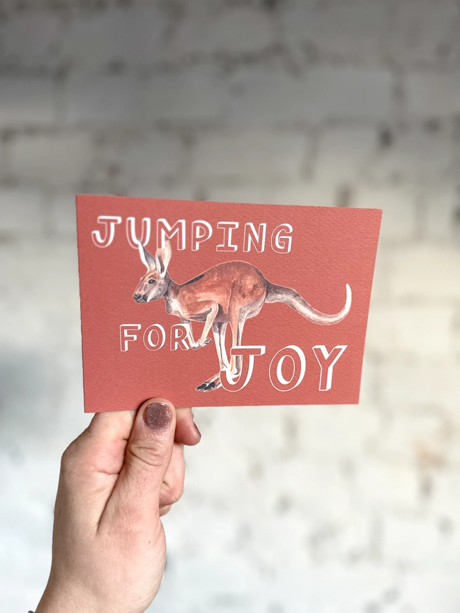 a rusty red greetings card featuring an illustration of a kangaroo with the text “jumping for joy” with a muted purple envelope