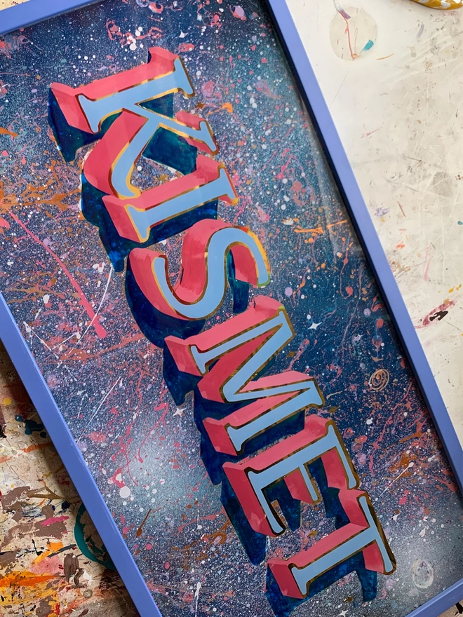 Gilded outlined letters spelling 'kismet' with a baby blue letter fill and pink shade, with splattered space-inspired background in a blue frame.