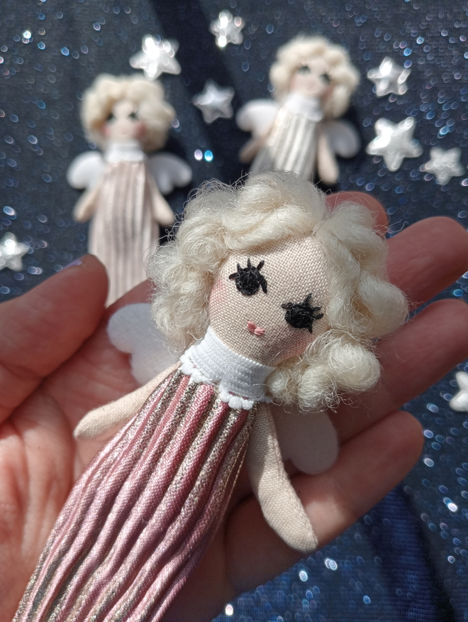 tiny angel doll nestled in the palm of a hand. doll is wearing a dusky pink gown with frilled collar. she has softly curled platinum white hair