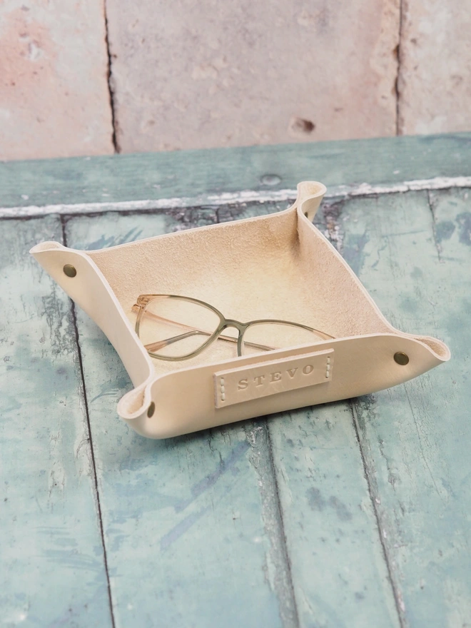 Personalised leather valet tray. A natural cream square leather tray, with a personalised name tag on one side. The tray holds a pair of reading glasses.