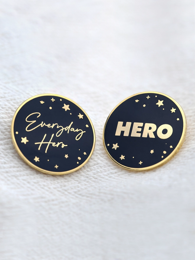 Two navy blue and gold enamel pin badges, each with a starry design and the words "Everyday Hero" on one and "Hero" on the other are resting on site fabric.