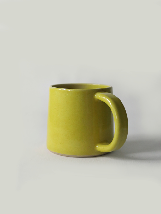 A handmade ceramic standard mug in glossy sun yellow glaze with a chunky loop handle, photographed against a plain light grey background