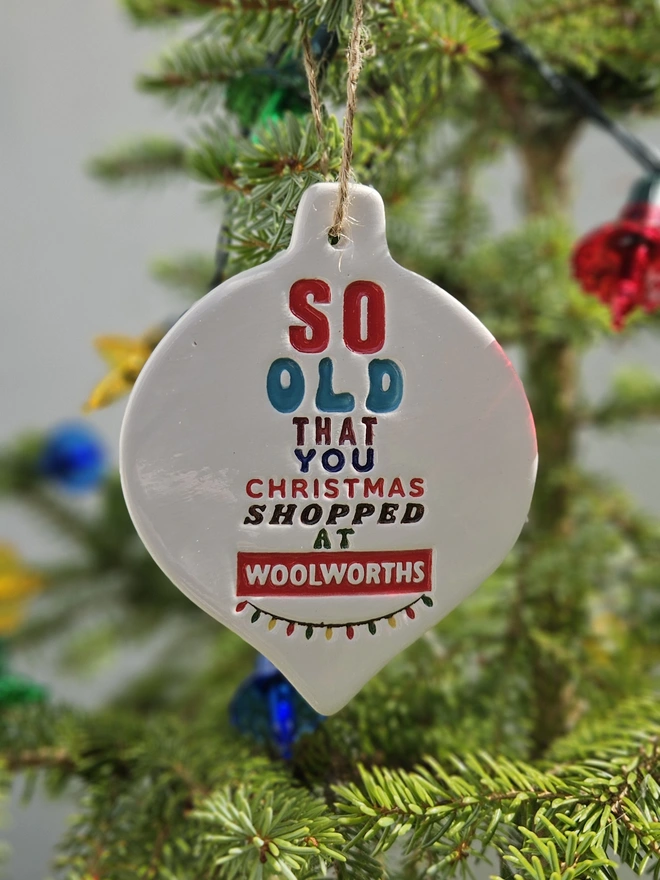 A handmade flat white ceramic bauble which measures 10cm  by 8cm with So old that you Christmas shopped at Woolworths written on it in colourful glazes and retro fonts with a little string of colourful fairy lights stamped into the clay.