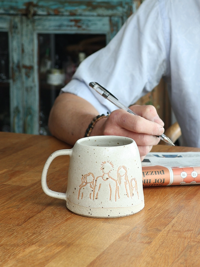 Childrens Drawing Handmade Ceramic Mug on a wooden table with a man writing on a newspaper in the background
