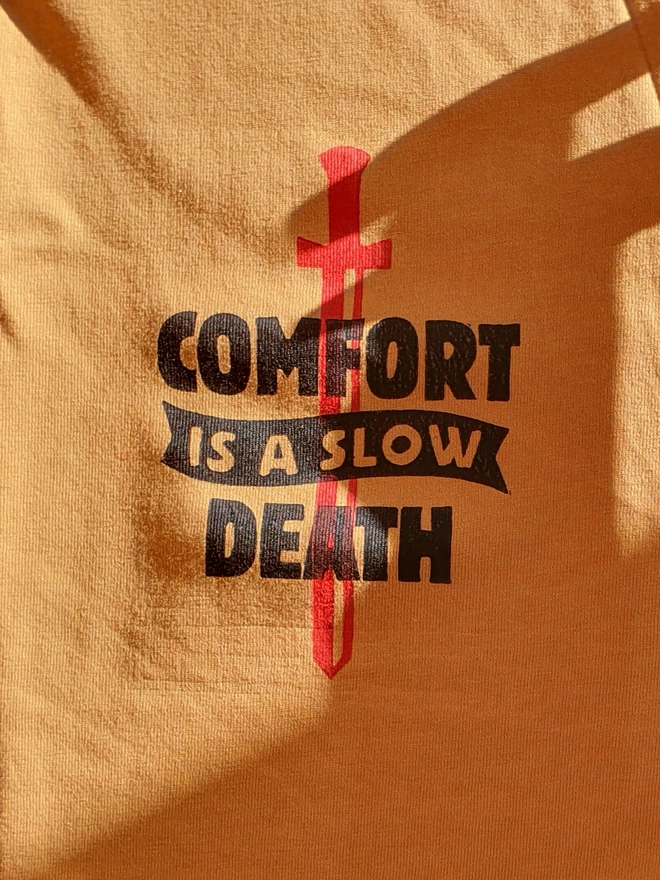 A yellow t-shirt with a black handprinted design on the front depicting a dagger and the phrase 'comfort is a slow death'