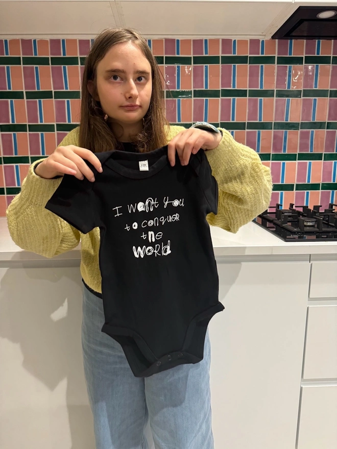 Piper the artist holds up the black baby grow / body suit with her white text on the front