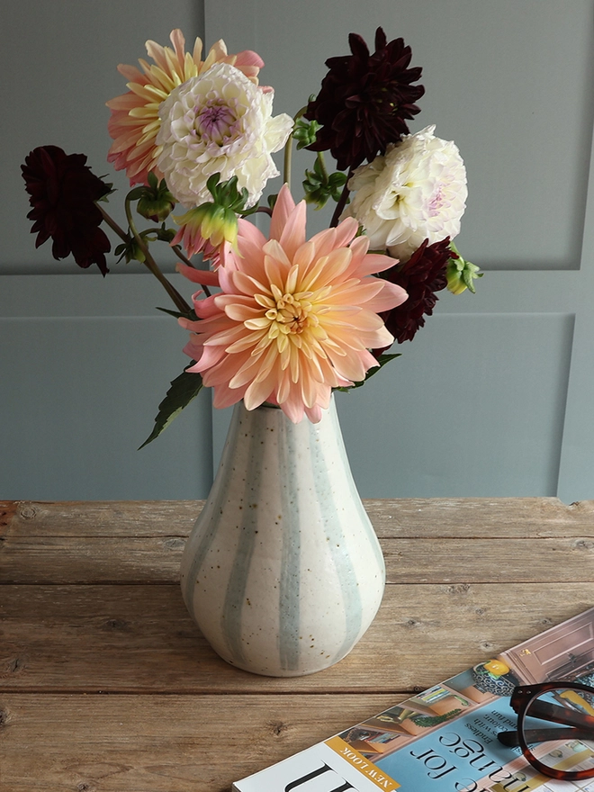 Large Handmade Ceramic Stoneware Vase in Blue and White Stripe holding a bouquet of pink, yellow and red flowers. The vase sits on a wooden surface next to a handmade ceramic white mug sitting on a ceramic coaster, and a magazine with a pair of glasses on top. 
