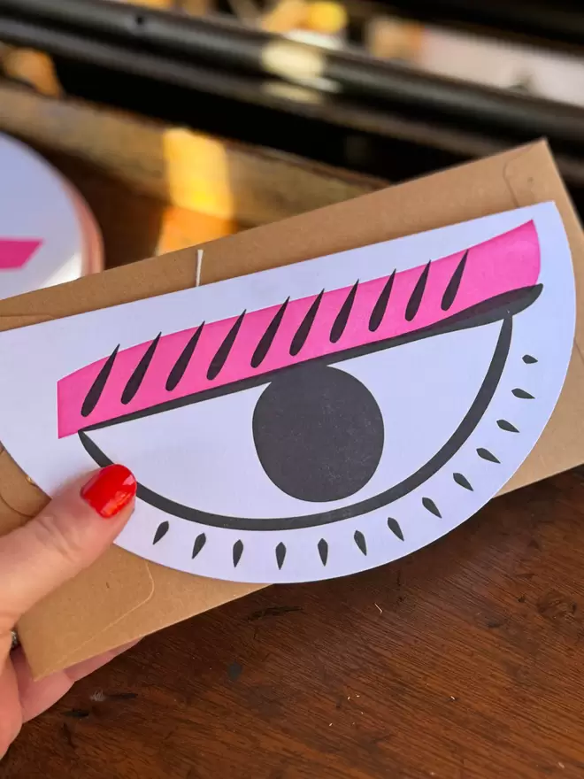 Lady holding half moon shaped card with large letterpress printed eye and bright pink eyeliner.