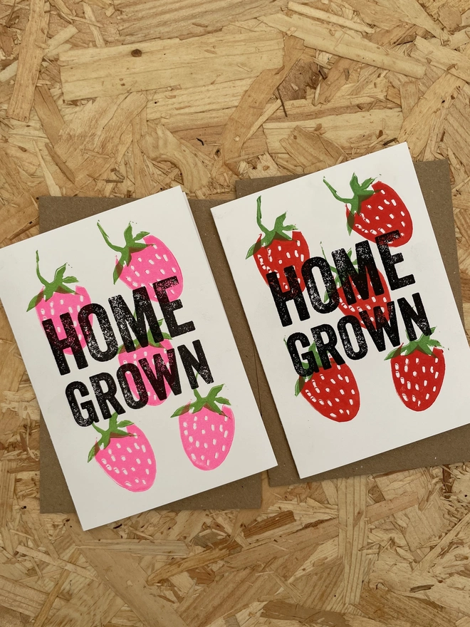 A white greeting card on a wooden surface shows  illustrated red and pink strawberries with green leaves surrounding bold black text that reads “HOME GROWN.”